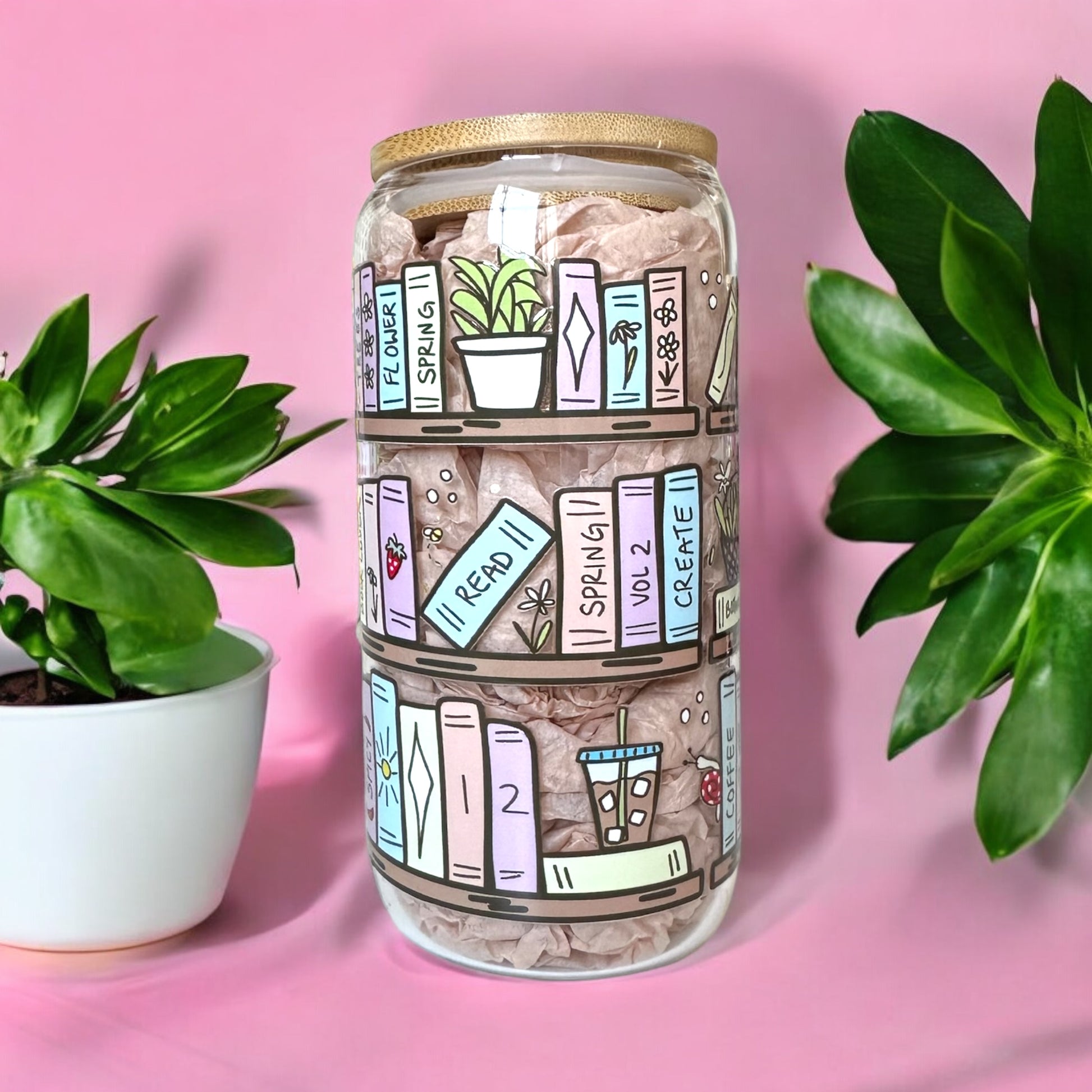 Decorative iced coffee glass cup with book-themed design on a pink background, surrounded by green potted plants.