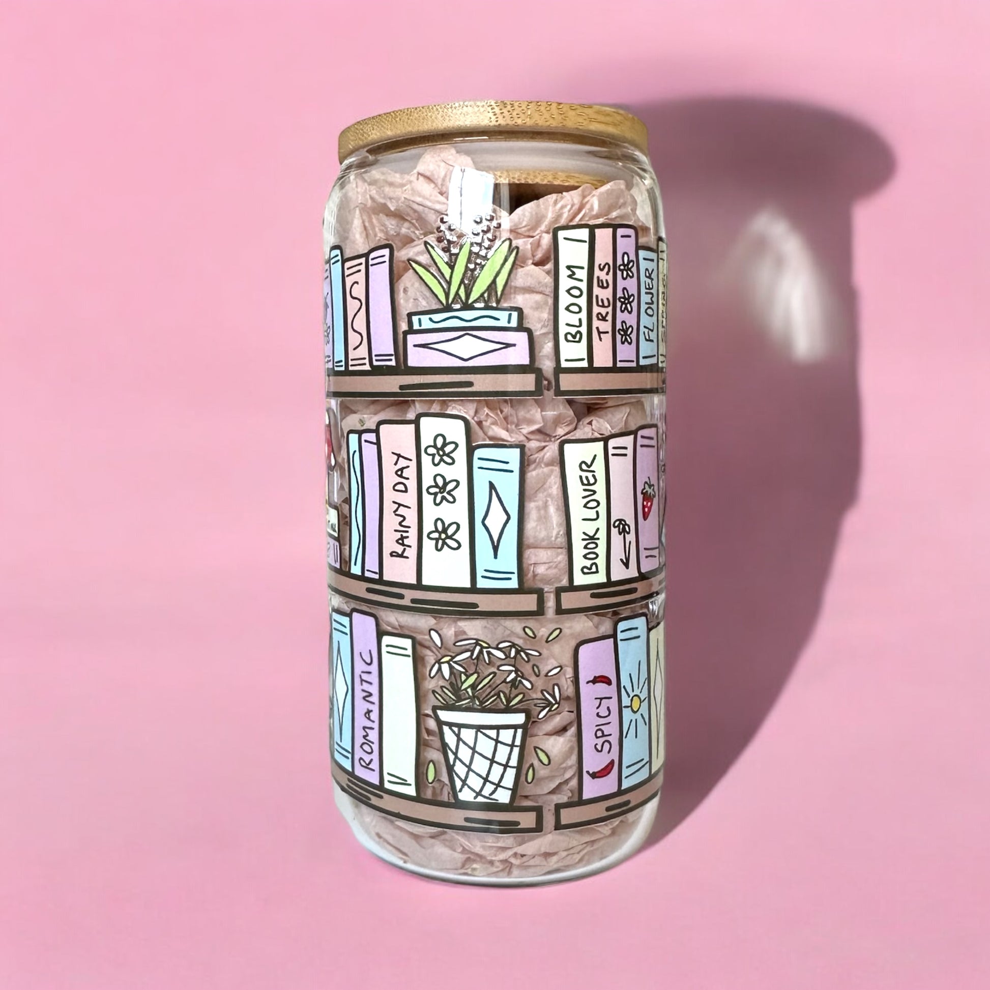 Decorative iced coffee glass cup featuring hand-drawn bookshelf design with plants and books on a pink background.